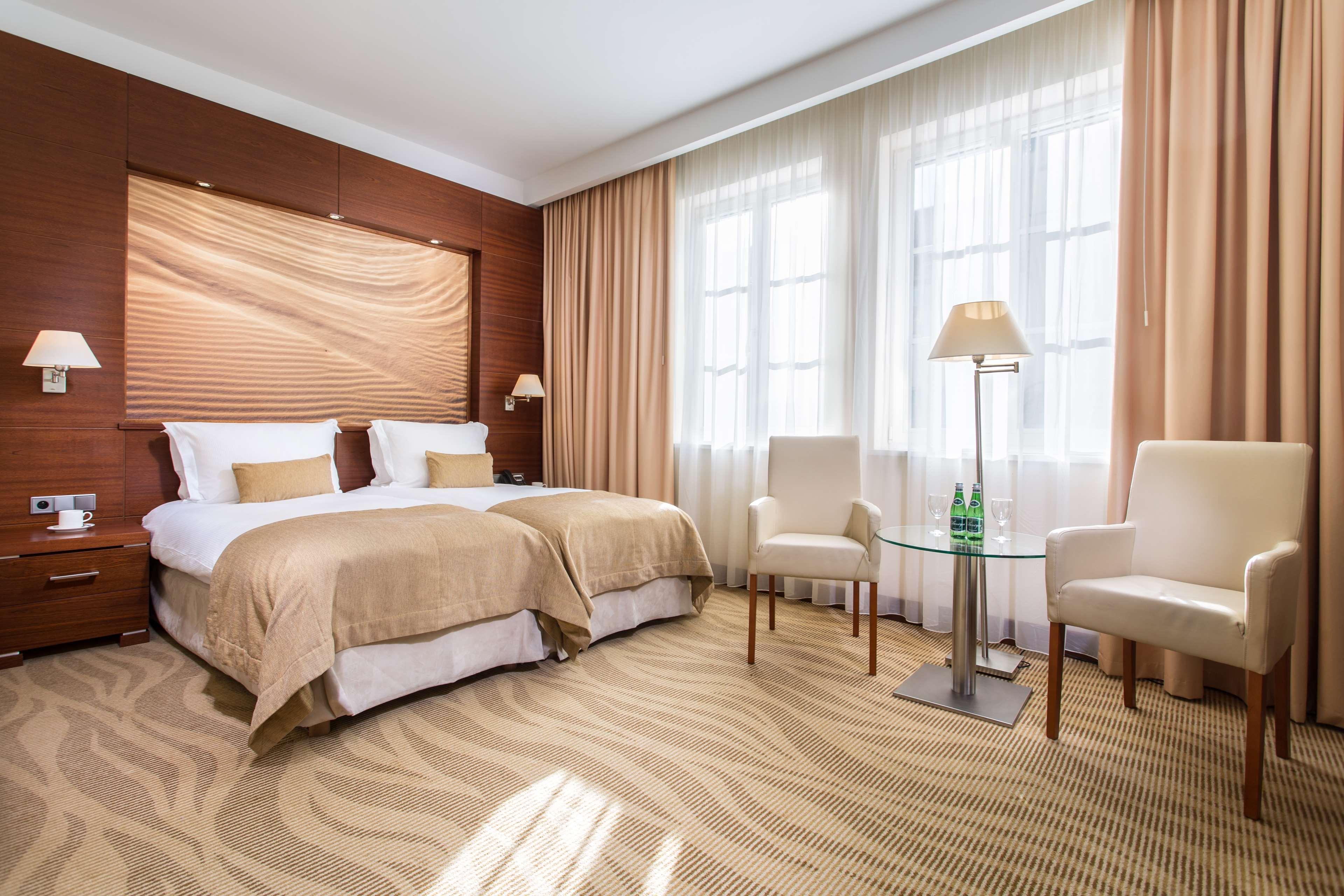 Radisson Blu Hotel, กดัญสก์ ภายนอก รูปภาพ The photo shows a well-decorated hotel room featuring two twin beds with beige linens and pillows. The walls have a warm, wooden paneling accent, and large windows are dressed with light curtains allowing natural light to fill the space. There are tw