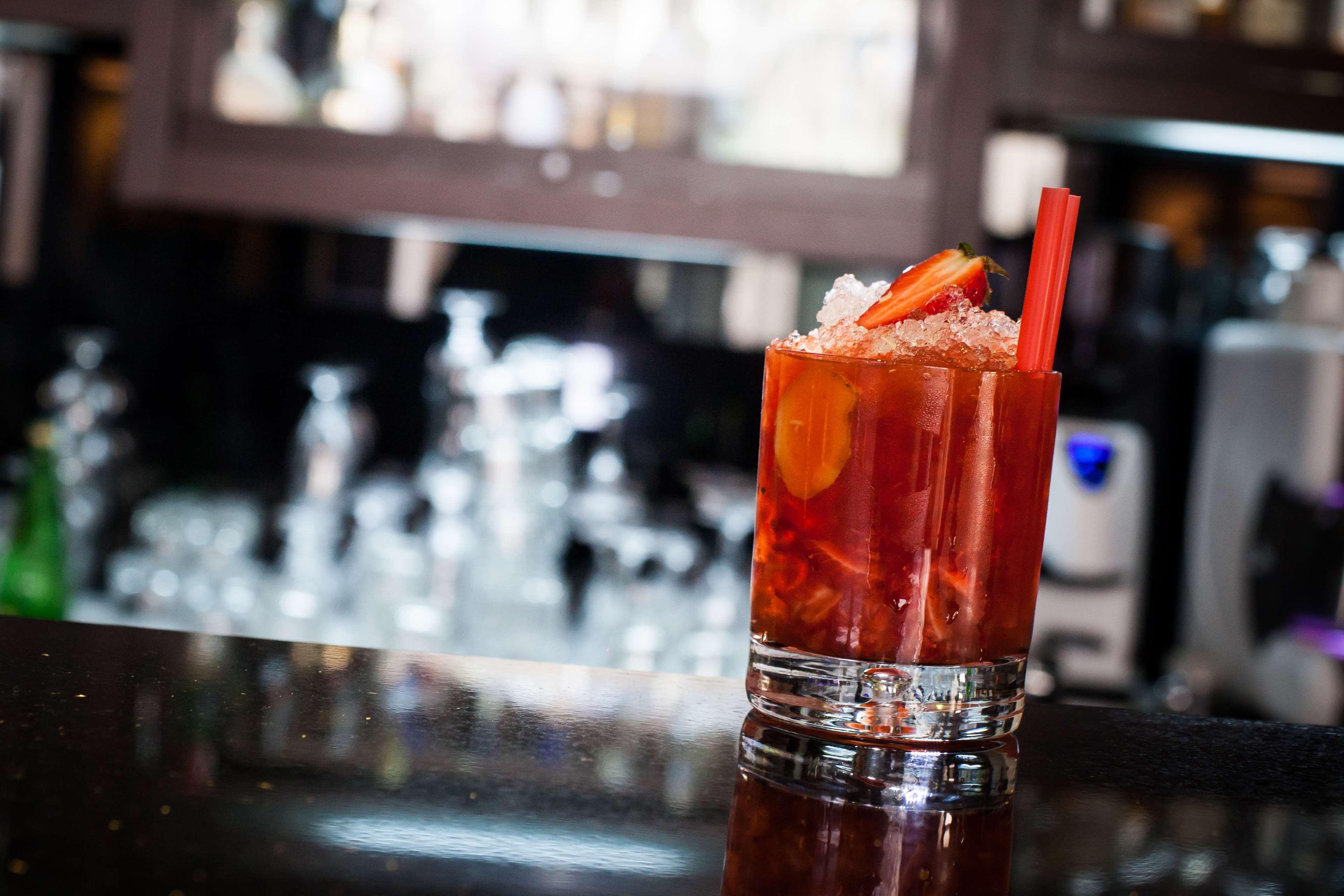 Radisson Blu Hotel, กดัญสก์ ภายนอก รูปภาพ The photo shows a glass of a colorful cocktail placed on a dark surface, likely a bar countertop. The drink is garnished with slices of fruit, possibly strawberries, and is filled with ice. In the background, there are blurred outlines of other glass