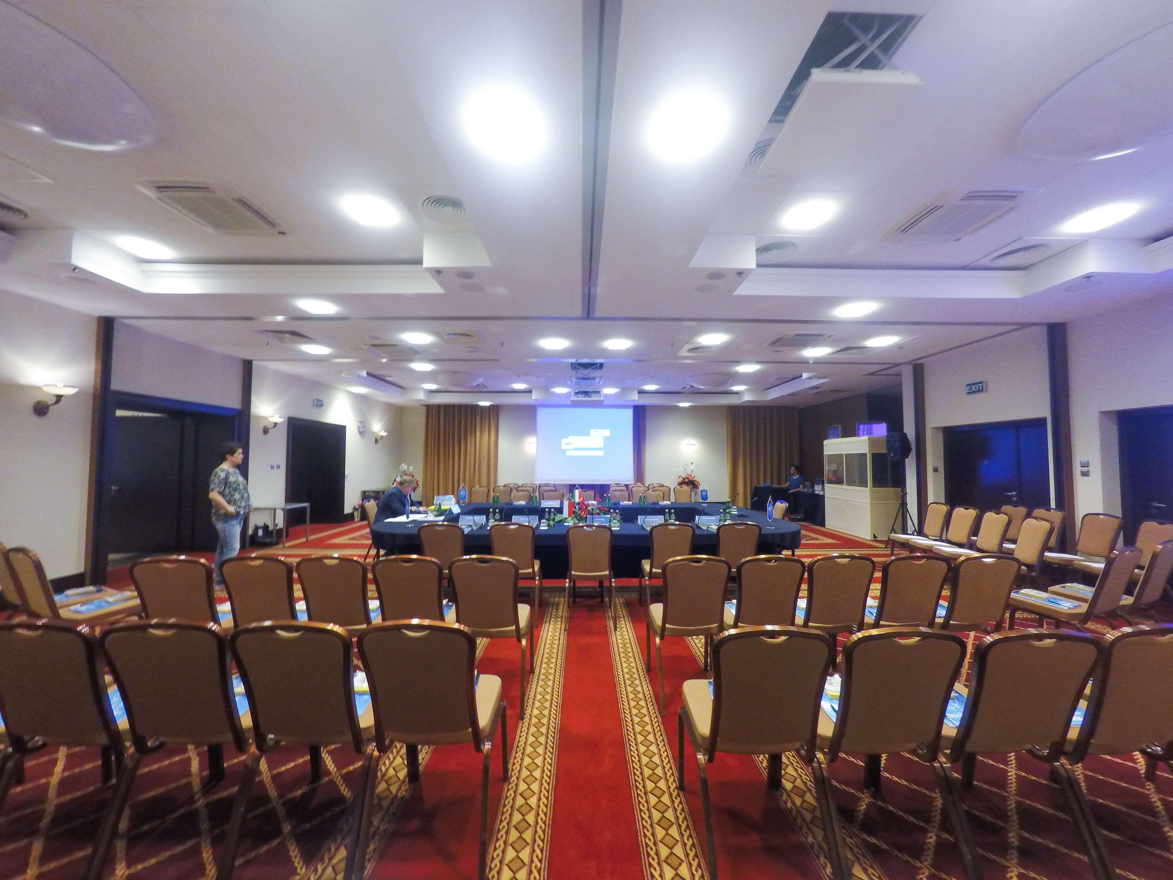 Radisson Blu Hotel, กดัญสก์ ภายนอก รูปภาพ The photo shows a conference room set up for a meeting or presentation. There are several rows of beige chairs arranged in front of a long table, which is also set up for participants. A large screen or projector is positioned at the front of the roo