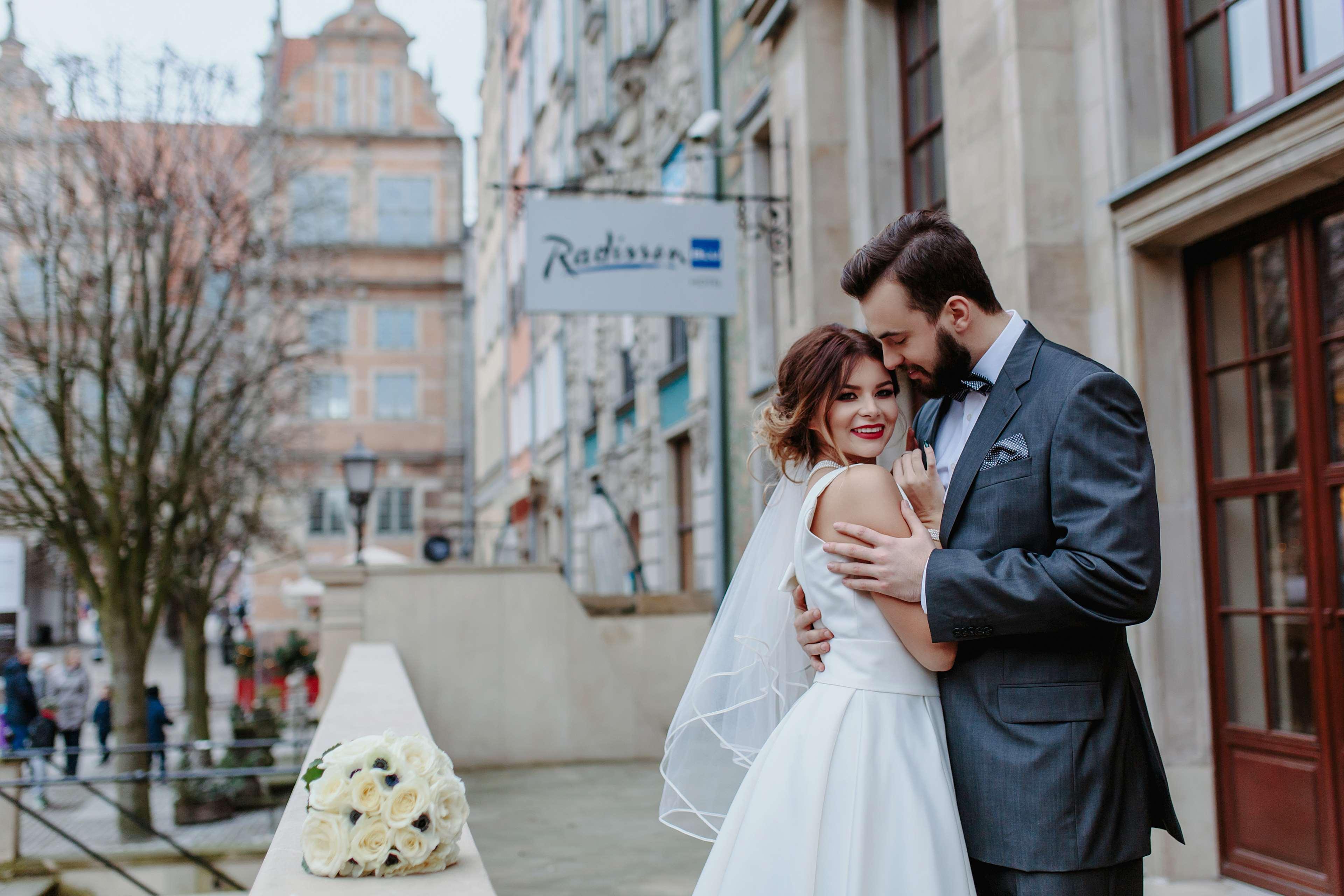 Radisson Blu Hotel, กดัญสก์ ภายนอก รูปภาพ The photo depicts a bride and groom embracing on a balcony. The bride is wearing a white wedding dress with a veil, and she is smiling while leaning into the groom. He is dressed in a grey suit and is holding her close, both appearing very happy. In 