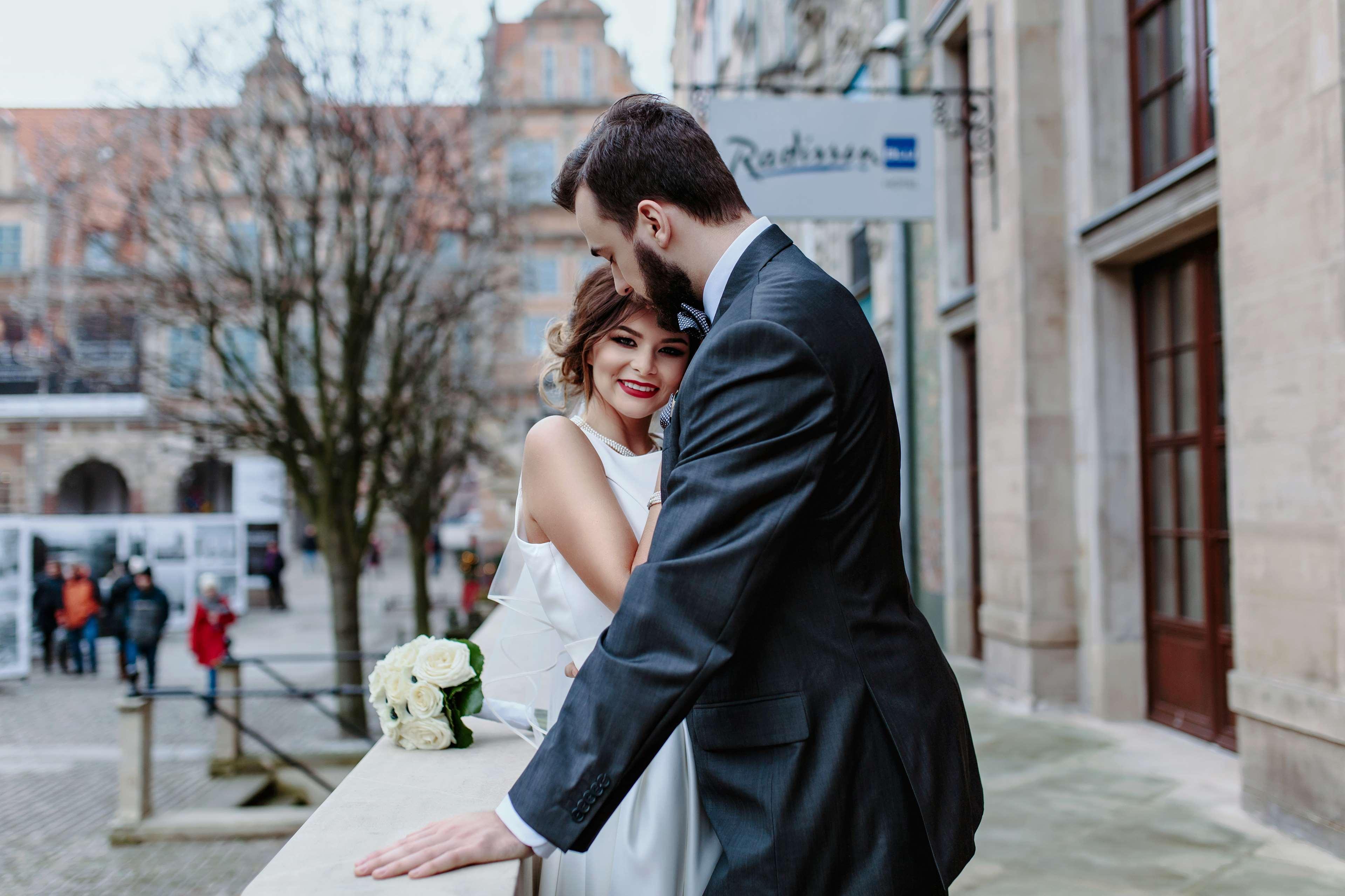 Radisson Blu Hotel, กดัญสก์ ภายนอก รูปภาพ The photo shows a couple embracing in an outdoor setting. The woman, dressed in a white wedding gown, is smiling and appears to be enjoying the moment. The man, wearing a dark suit, leans in toward her, suggesting a romantic atmosphere. In the backgr
