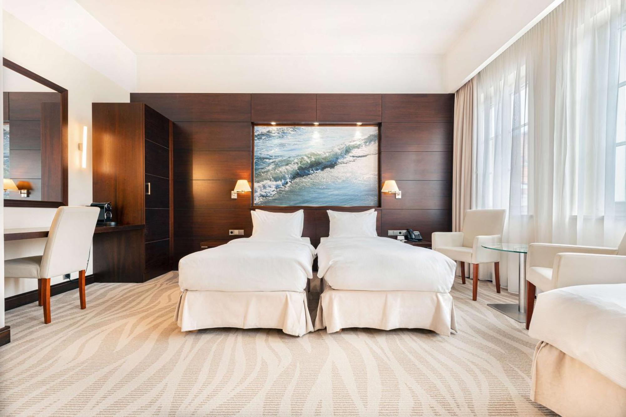 Radisson Blu Hotel, กดัญสก์ ภายนอก รูปภาพ The photo shows a modern hotel room featuring two twin beds dressed in white bedding. The room has a warm, elegant design with wooden cabinetry on one wall and a large piece of artwork depicting a landscape or seascape above the beds. There are two b