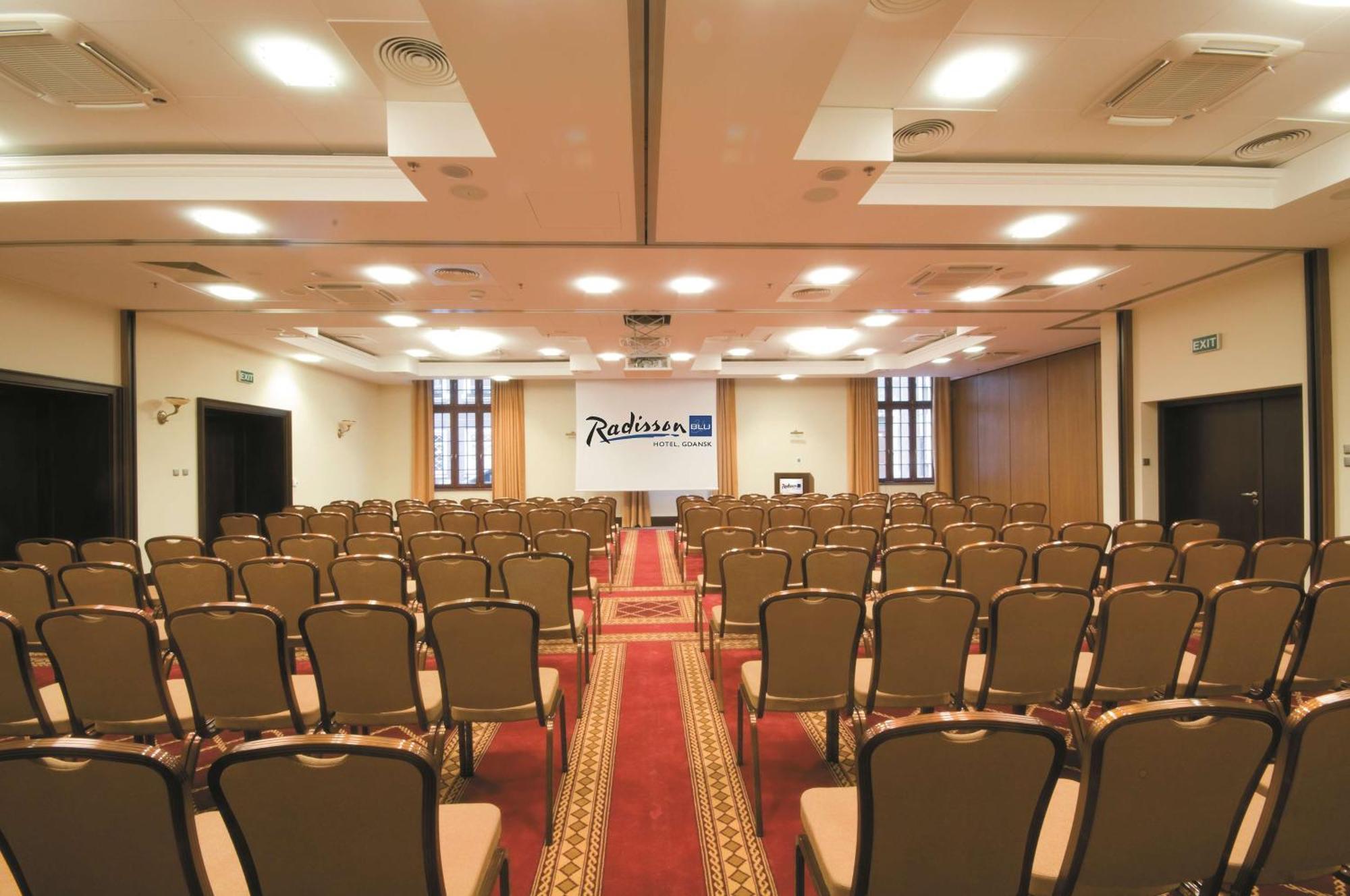 Radisson Blu Hotel, กดัญสก์ ภายนอก รูปภาพ The photo shows the interior of a conference room or event space. It features rows of brown chairs arranged neatly, facing towards the front where a stage or presentation area is set up. There is a carpet running down the center of the room, and the 