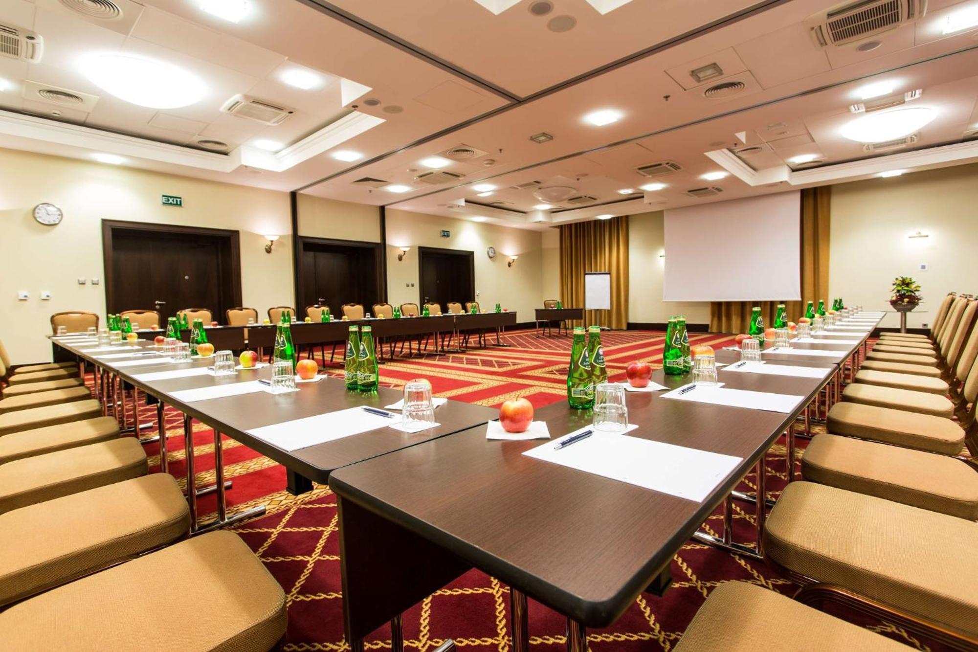 Radisson Blu Hotel, กดัญสก์ ภายนอก รูปภาพ The photo shows a conference room set up for a meeting or seminar. Long tables are arranged in a U-shape, surrounded by beige chairs. Each table has green glass bottles of water, small glasses, and apples placed on it. There are also sheets of paper 