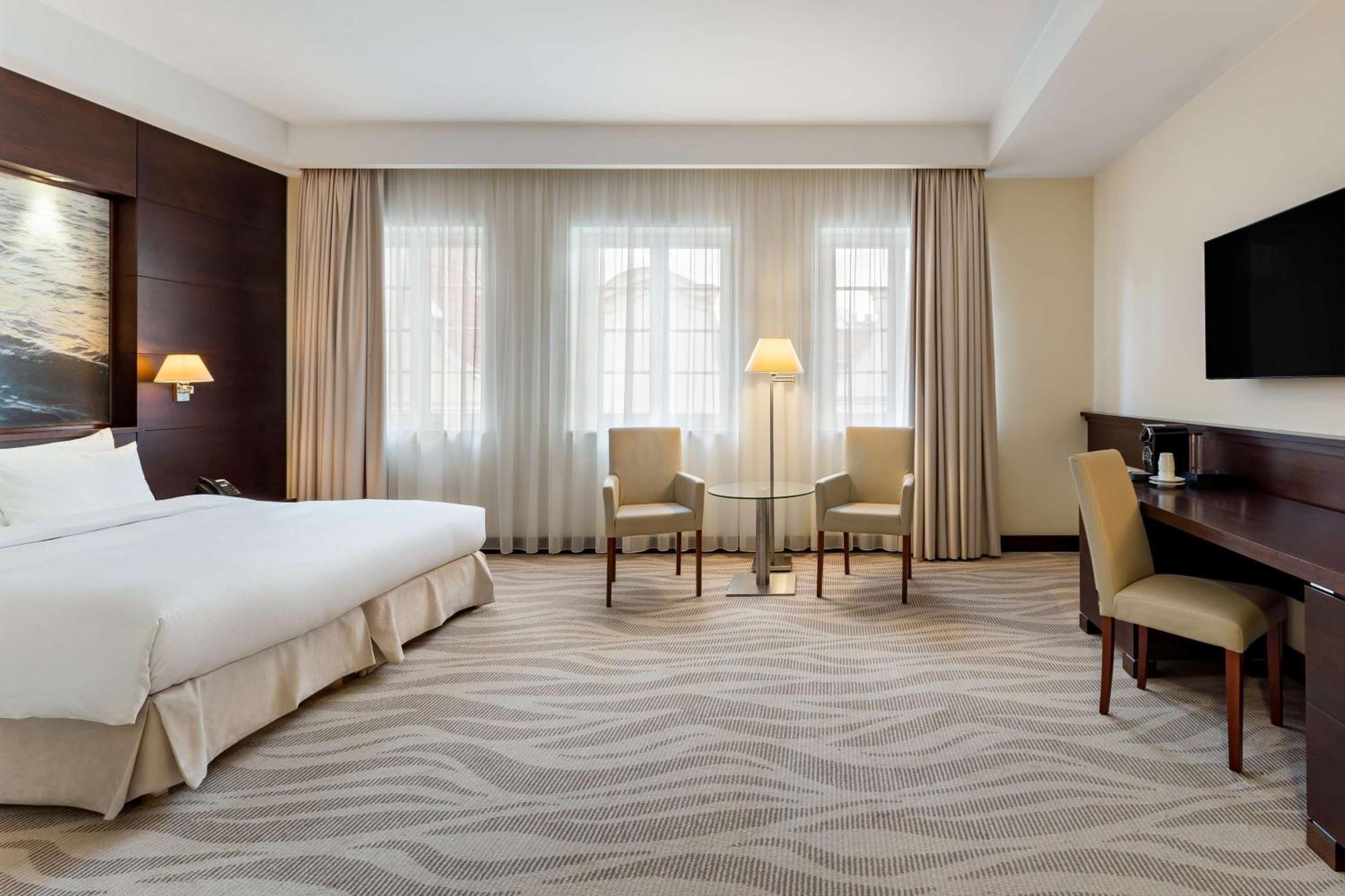 Radisson Blu Hotel, กดัญสก์ ภายนอก รูปภาพ The photo shows a hotel room featuring a modern and clean design. There's a large bed with white bedding positioned against a wall, accompanied by bedside tables. Two armchairs and a small side table are set near a lamp in front of large windows that