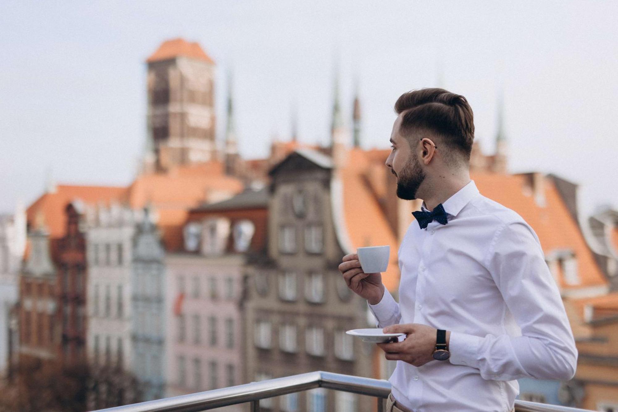 Radisson Blu Hotel, กดัญสก์ ภายนอก รูปภาพ The photo features a man standing on a balcony, holding a cup of coffee or tea in one hand while resting a saucer in the other. He is dressed in a white shirt with a bow tie. The background reveals a picturesque view of an old town, showcasing buildi